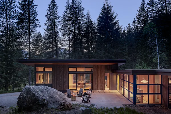 Dusk view of a modern hillside house with wood siding, floor-to-ceiling glazing, and a concrete patio with seating around a fire pit.