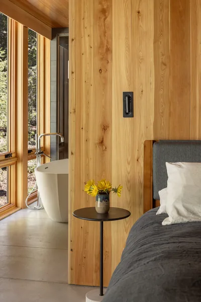 A bedroom corner with wood paneling and a side table next to a bathroom with a freestanding tub and floor-to-ceiling windows