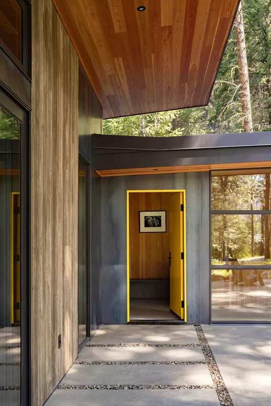 Modern house entrance features a yellow door framed by dark metal and wood paneling, with a concrete walkway accented by gravel.