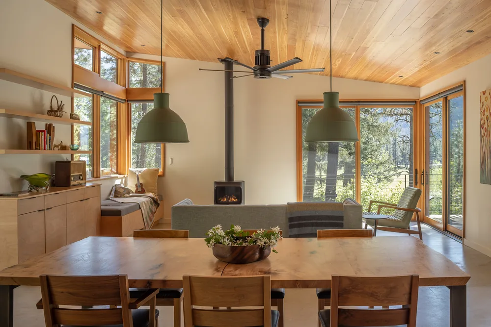 Open-plan living and dining room with wood-paneled ceiling, large windows, and a central wood stove.