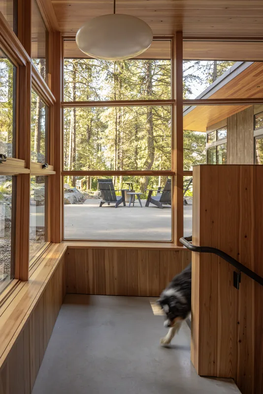 Entry area with large wood-framed windows looking out to a patio with Adirondack chairs and forested landscape.