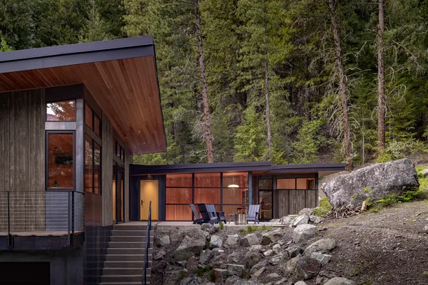 Modern hillside house exterior with dark wood cladding and floor-to-ceiling glazing in a forest setting.