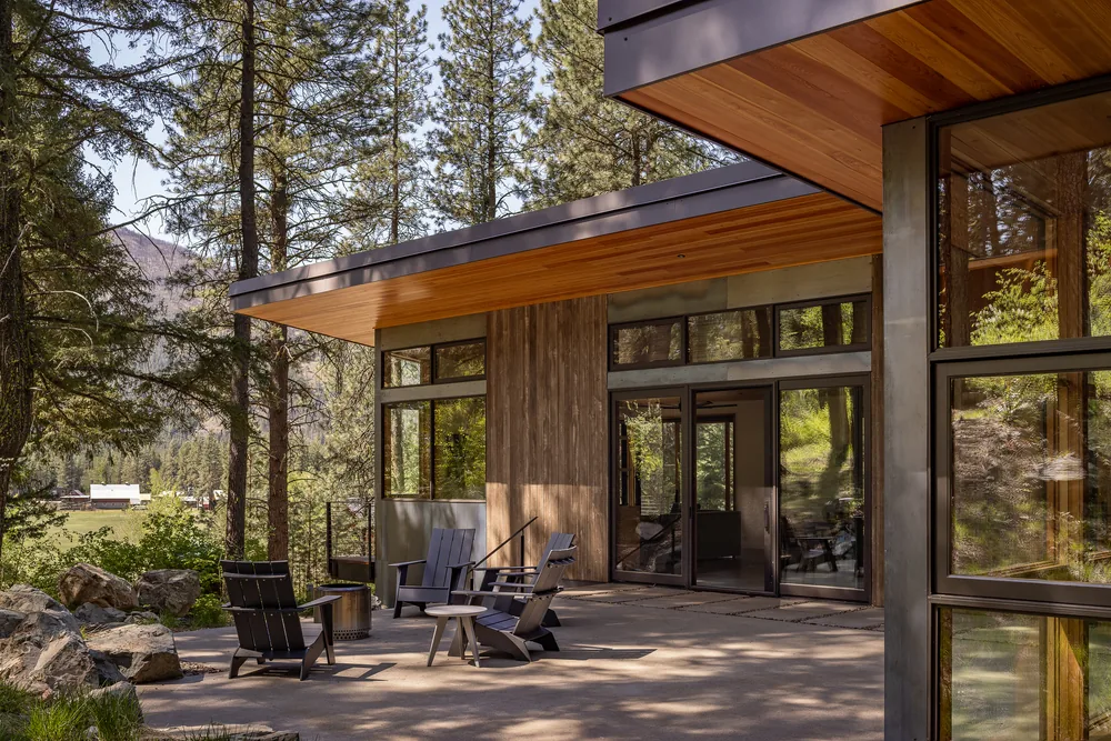 Concrete patio with outdoor seating at the rear of a modern hillside house featuring large windows and wood soffits.