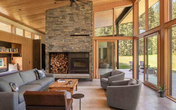 Living room features a stone fireplace, wood ceiling, and large windows opening to a patio with outdoor chairs.