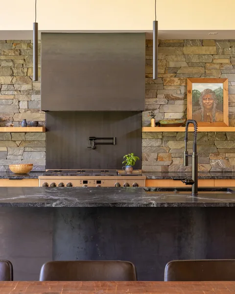 Kitchen features a stone wall with floating wood shelves, a black metal range hood, and a black waterfall island with integrated sink and faucet.