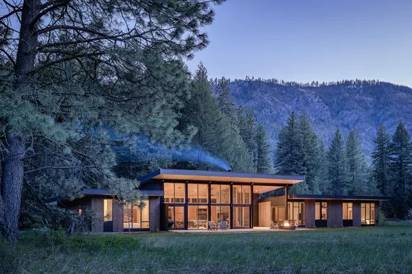 Modern single-story house with extensive floor-to-ceiling glazing set in a forested mountain landscape at dusk.