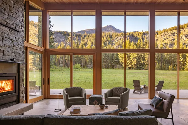 Living room with large wood-framed floor-to-ceiling windows and stone fireplace overlooking a meadow and mountains.