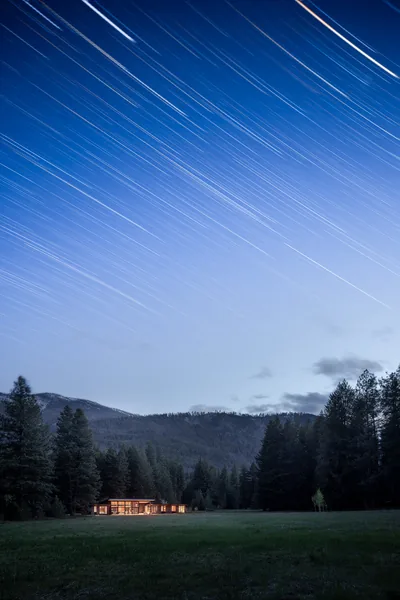 Night view of a modern house in a meadow with star trails visible in the sky above.