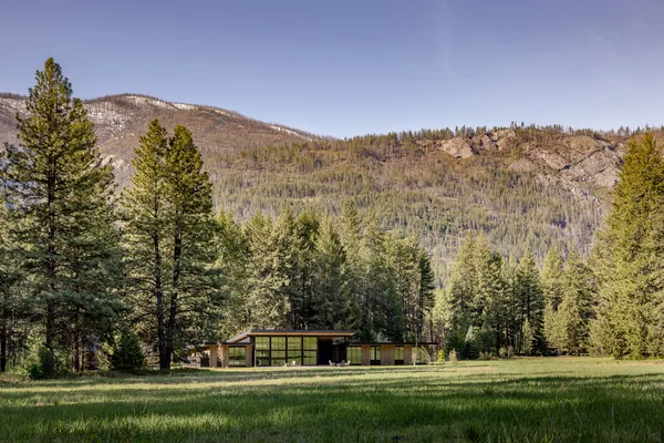 Modern house with floor-to-ceiling glazing located in a meadow surrounded by trees and mountains.