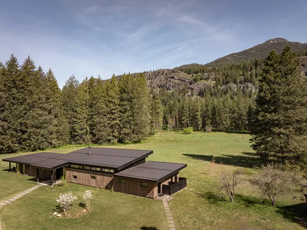 A flat-roofed wooden house in a meadow with forest and hills behind it.