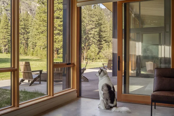 Interior looking through wood-framed glass windows and sliding door to outdoor patio with lounge chairs and a dog sitting inside.