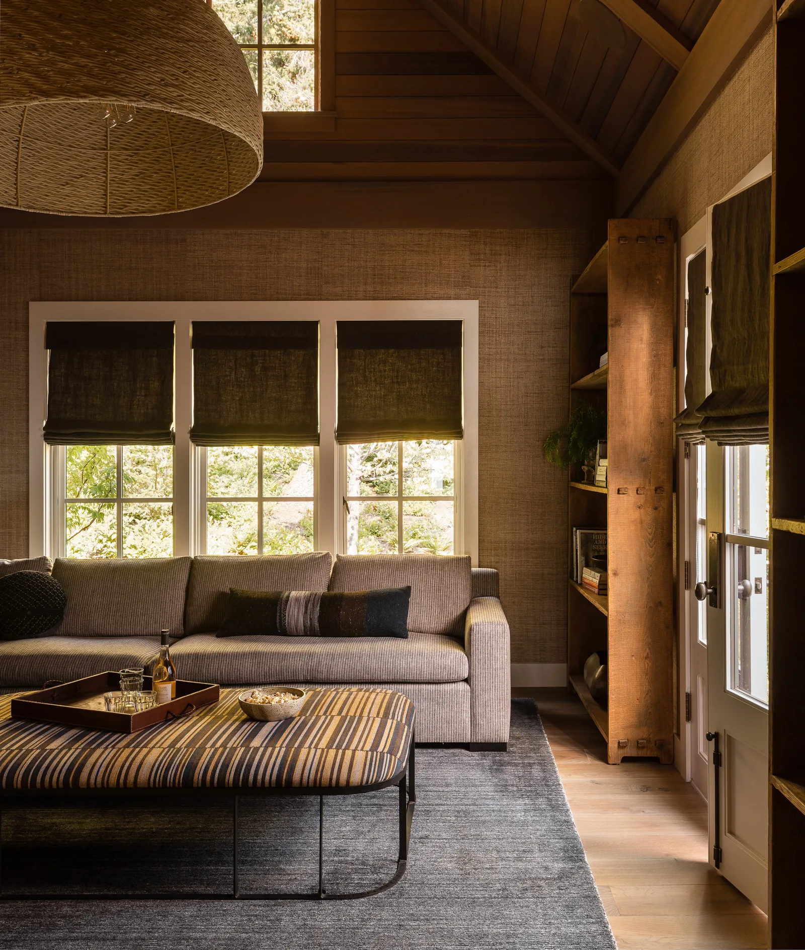 Living room featuring a grey sofa and striped ottoman with three windows and black roman shades, wood panel ceilings, and built-in wooden bookshelves.
