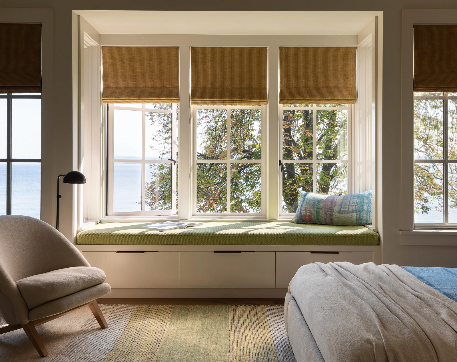 Bedroom interior showing a window seat with green cushion under four white-framed windows with woven shades partially lowered.