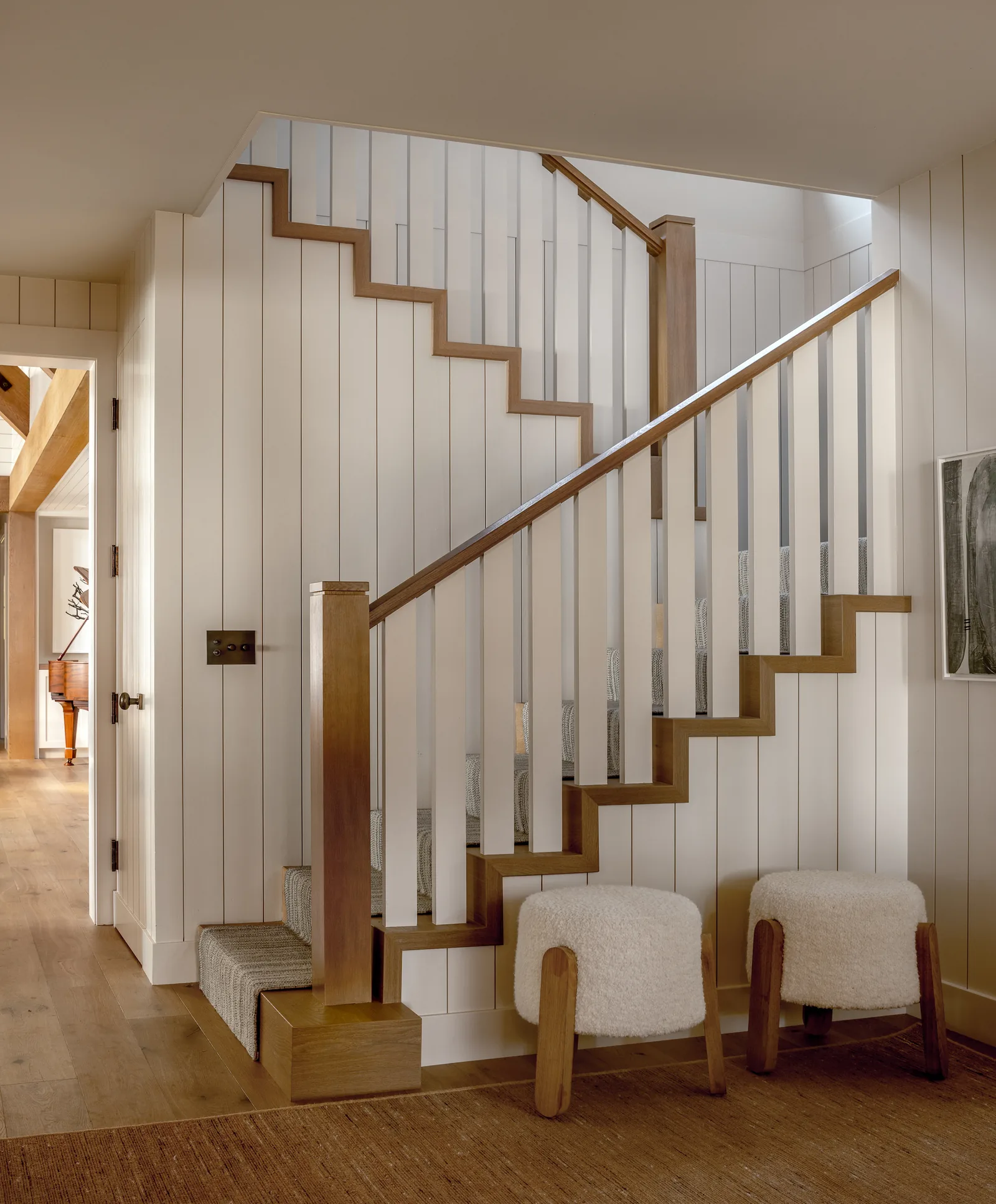 Interior view of a staircase with wooden handrails and white balustrade panels leading up with two small upholstered stools at the base.