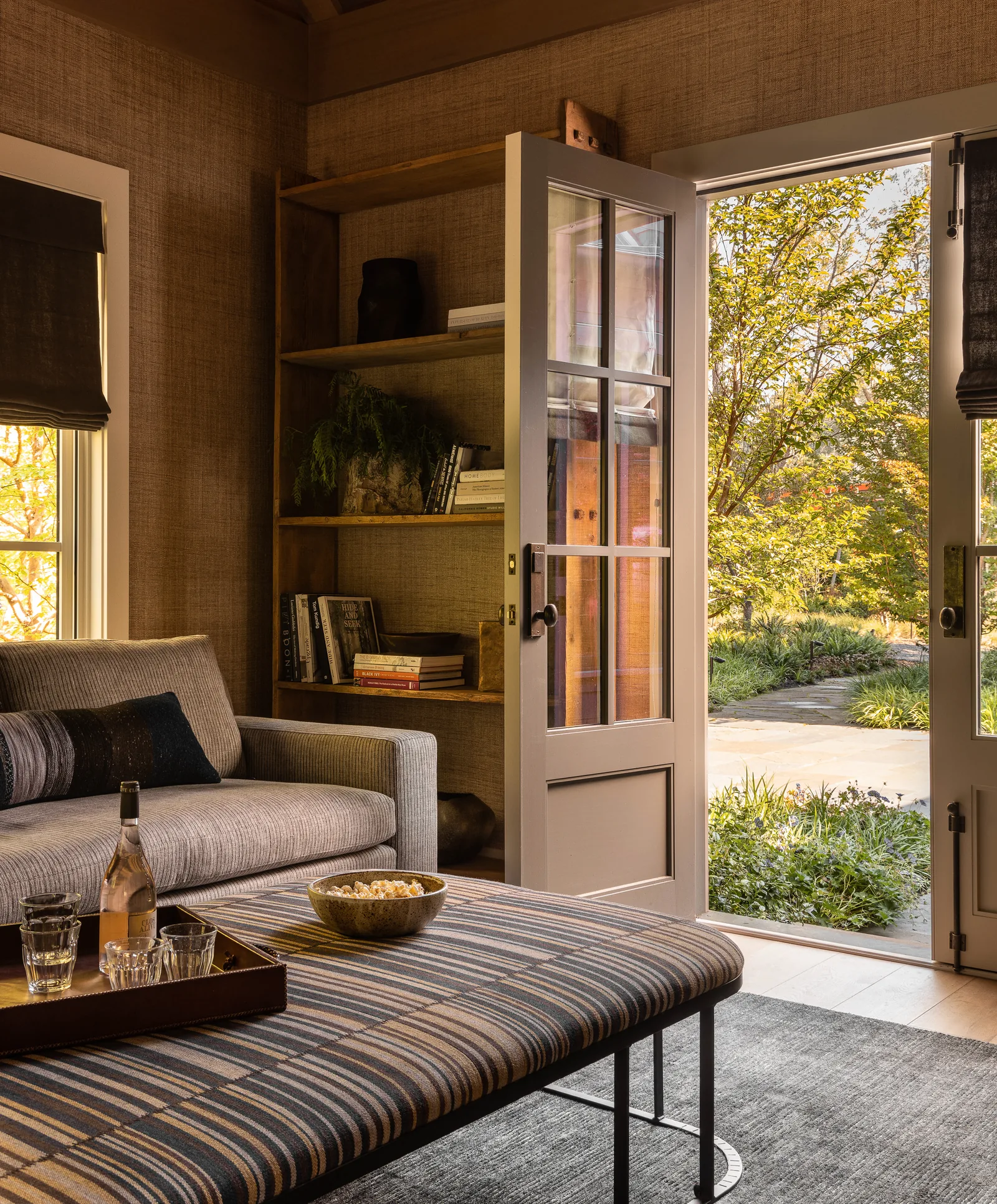 A living room interior with double glass doors opening onto a garden patio with plants and trees.