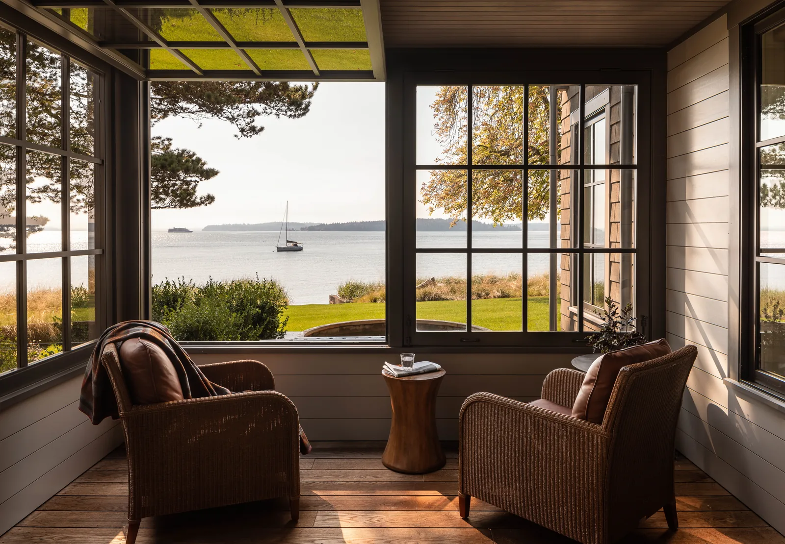 Porch seating area with large multi-pane windows framing a view of the lawn and waterfront with a sailboat.