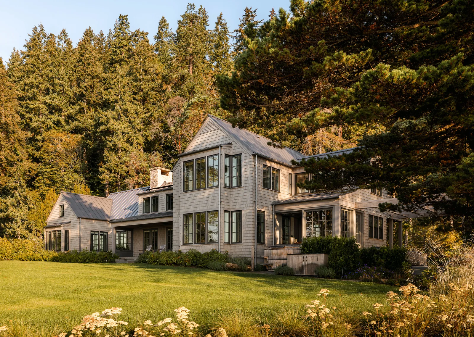 Exterior front view of a two-story house clad in wood shingles with multiple gabled metal roofs adjacent to a dense forest.