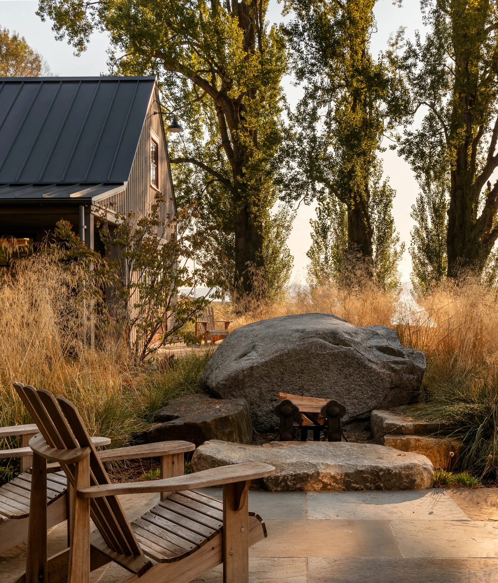 Outdoor patio featuring wooden Adirondack chairs and a stone fire pit next to a building with vertical siding and metal roof.