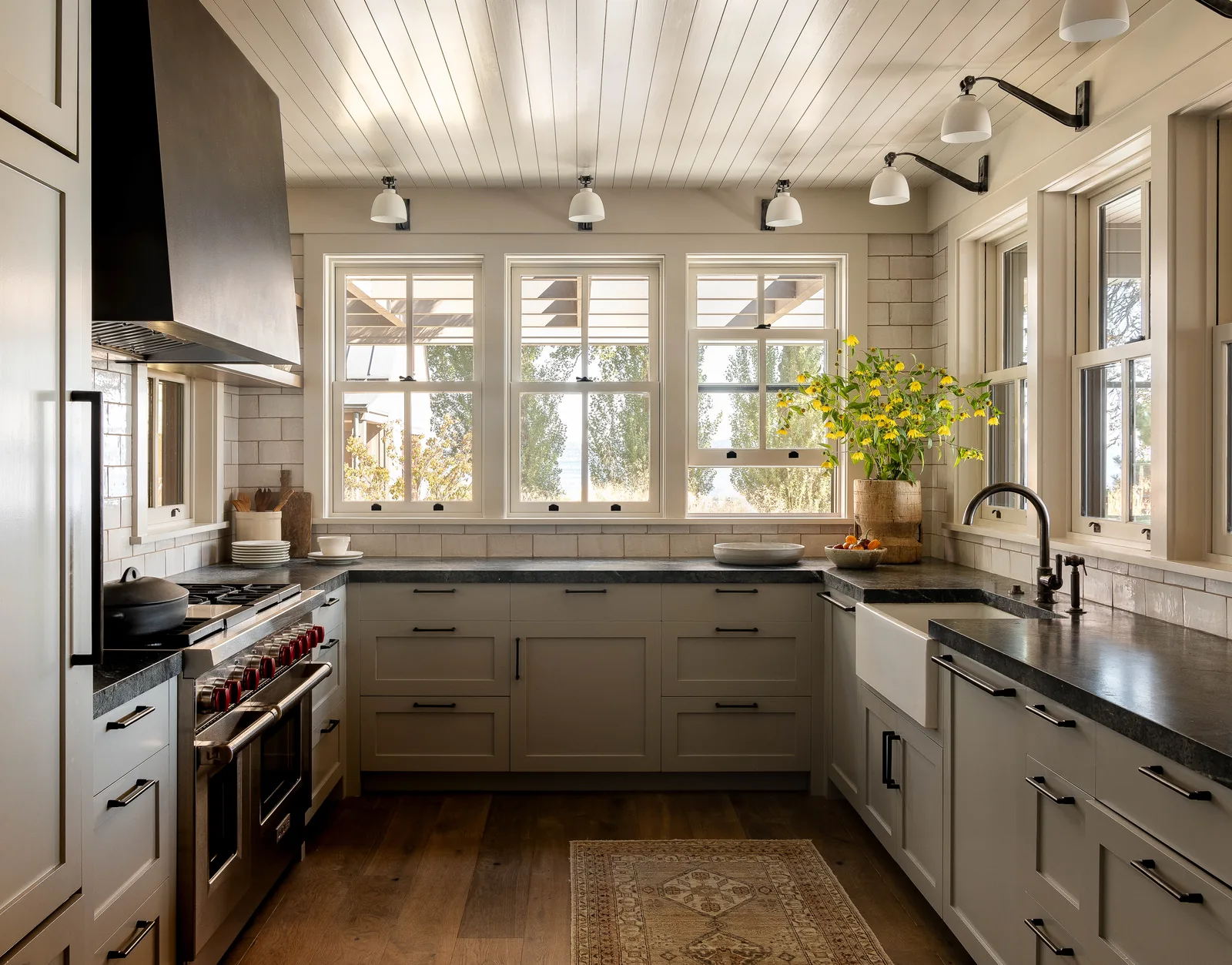 Kitchen featuring wood flooring, light cabinetry, black countertops, farmhouse sink, and four windows overlooking an exterior pergola.