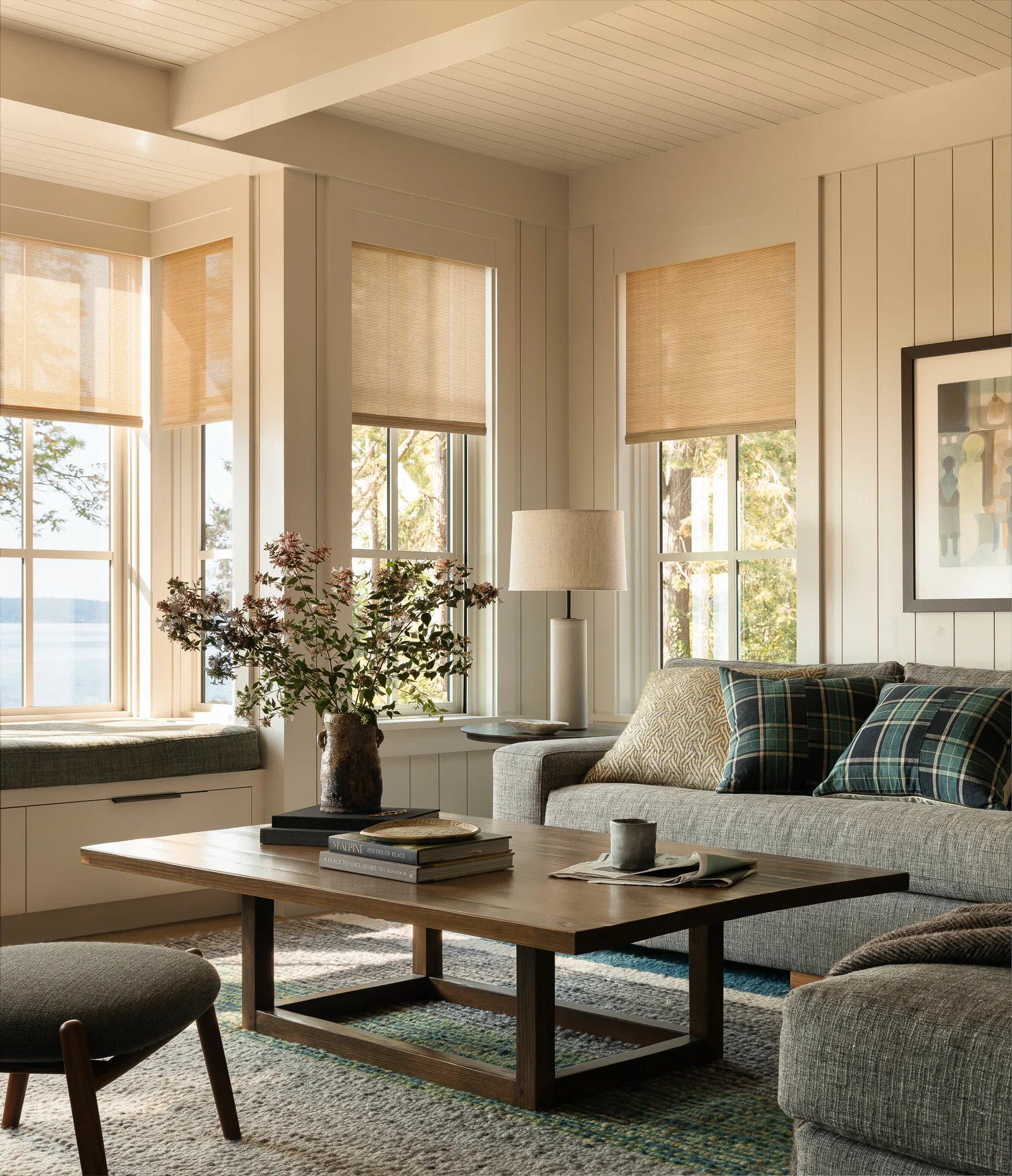 Living room features multiple windows with woven shades, a gray sofa with plaid pillows, wooden coffee table, and a built-in window seat.
