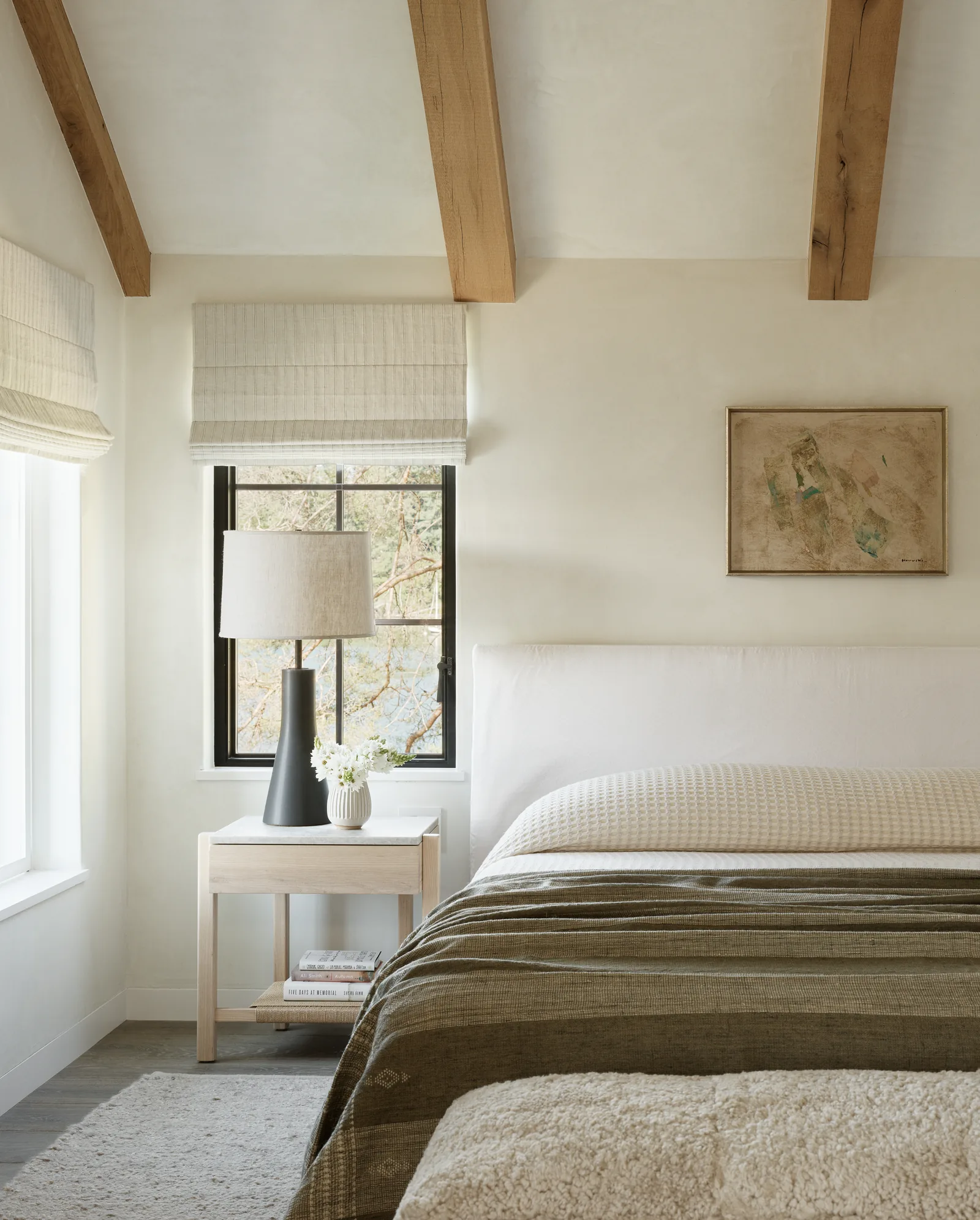 Bedroom interior featuring exposed wooden beams, a bed, a nightstand with a lamp and vase, and a window with a fabric roman shade.