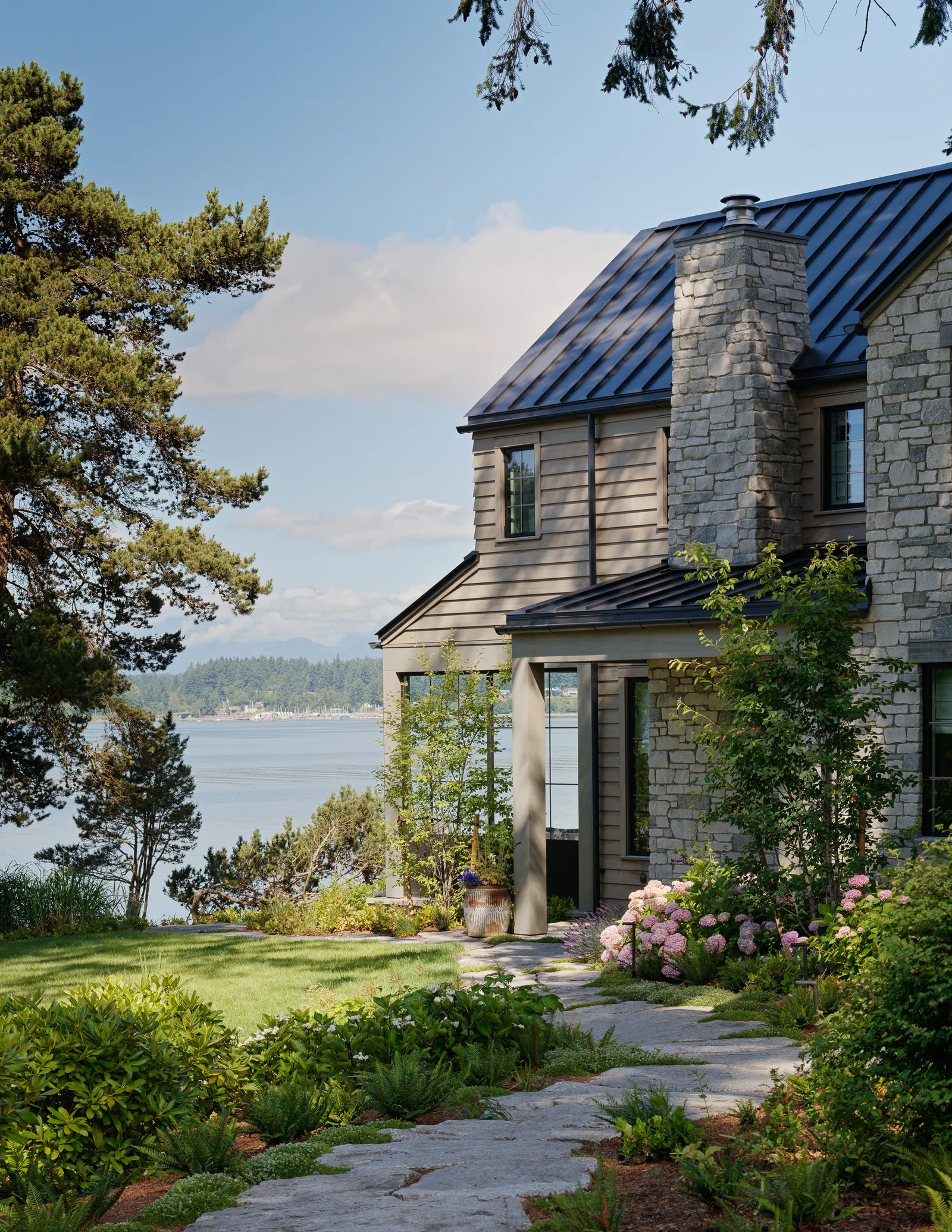Side exterior of home with stone chimney, metal roof, and landscaped garden path facing waterfront.