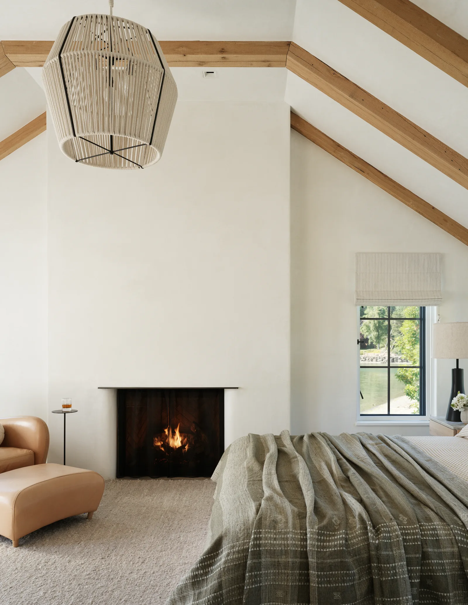 A bedroom with a fireplace, exposed wooden beams on a vaulted ceiling, and a window showing a water view.