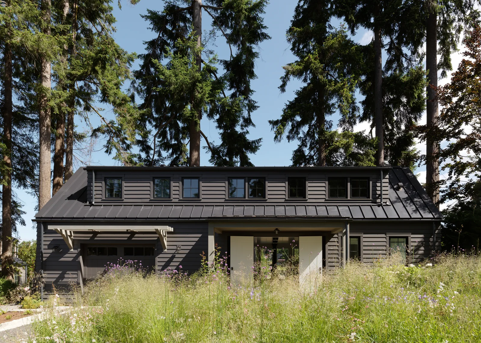 The Arrow Bay project features a modern barn-style house with a standing seam metal roof and multiple dormer windows on its front facade.