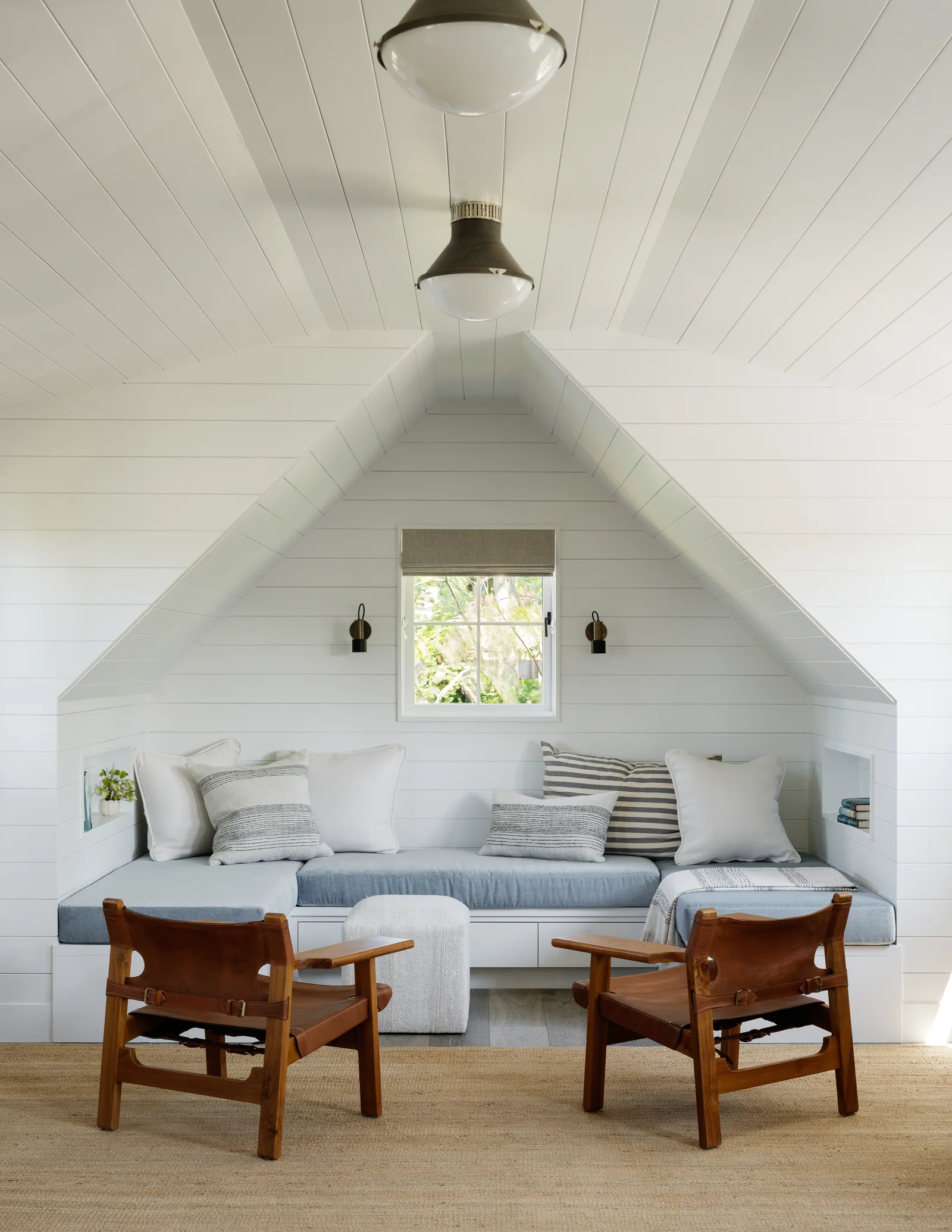 Seating nook with built-in bench under angled ceiling and paneled walls, furnished with wooden chairs and blue cushions.