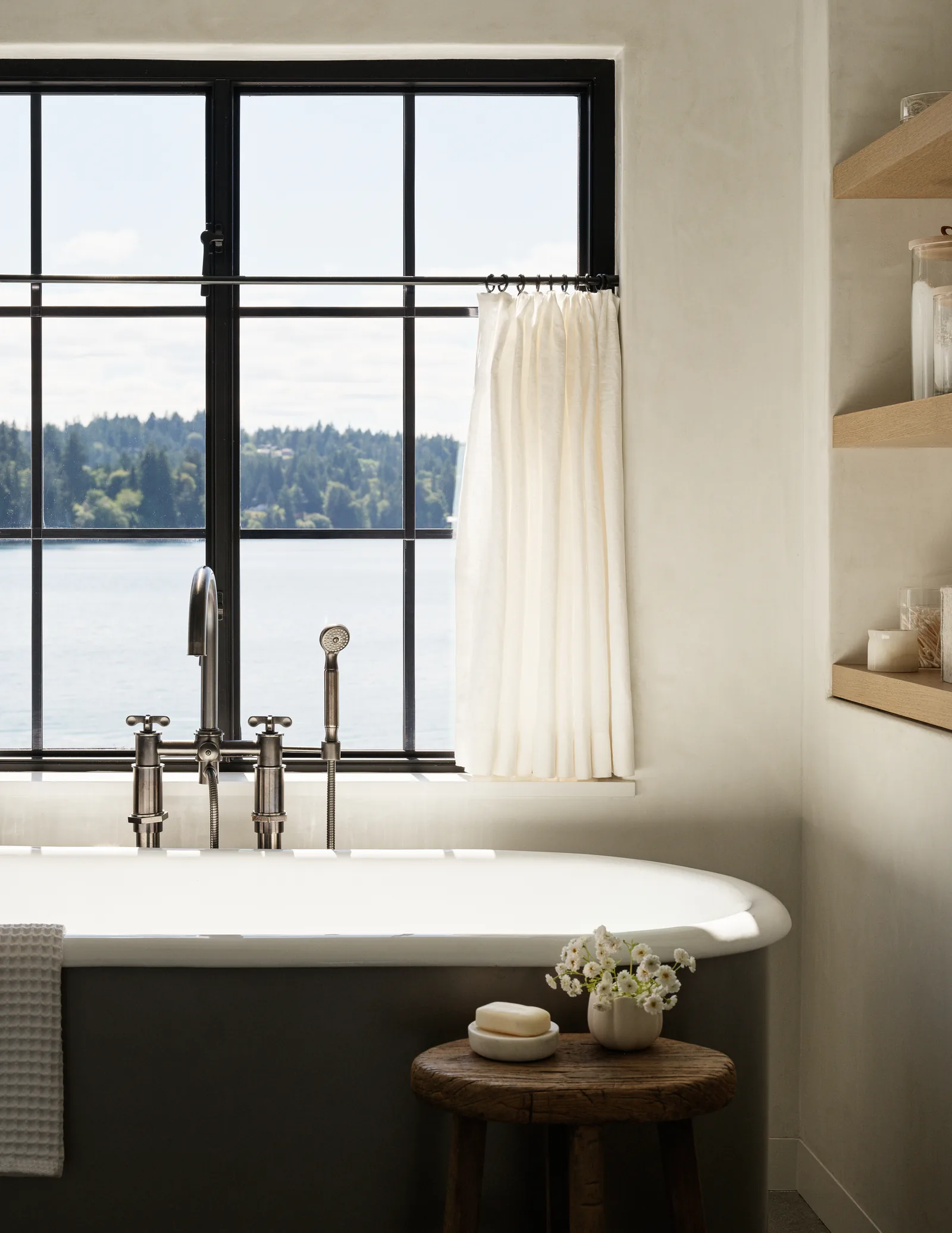 Bathtub and fixtures positioned beneath a divided light window with a white curtain in a bathroom with light-colored walls and wooden shelves.