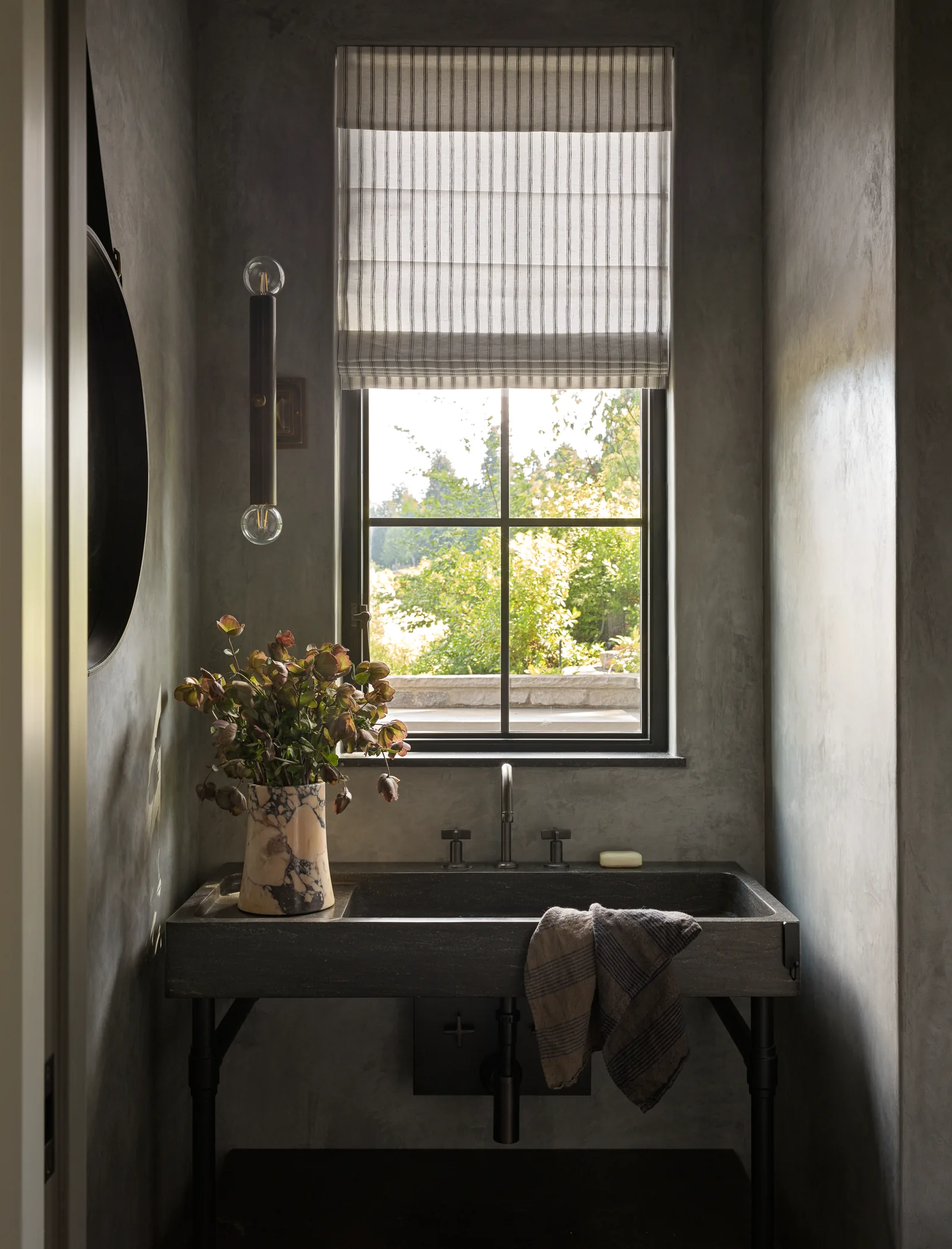 Bathroom with dark stone sink beneath a window with a striped Roman shade and a decorative vase with flowers.