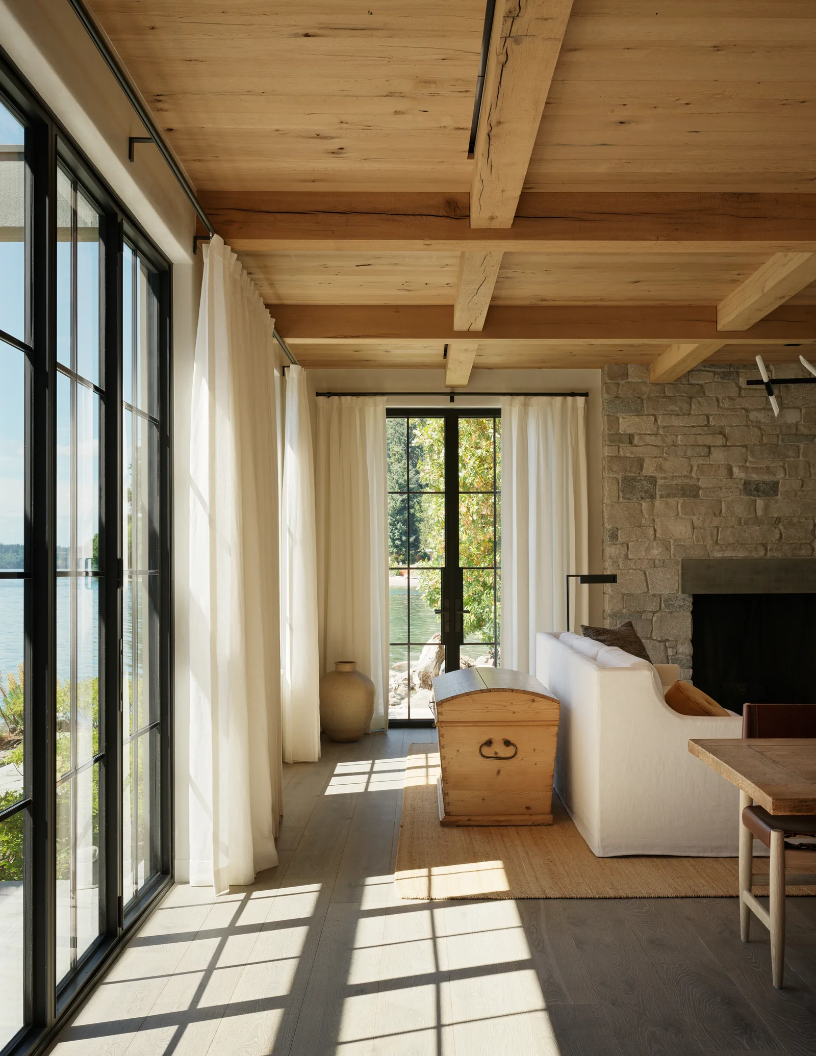 Living room with exposed wood ceiling beams, stone fireplace, and floor-to-ceiling black framed glass doors overlooking water.