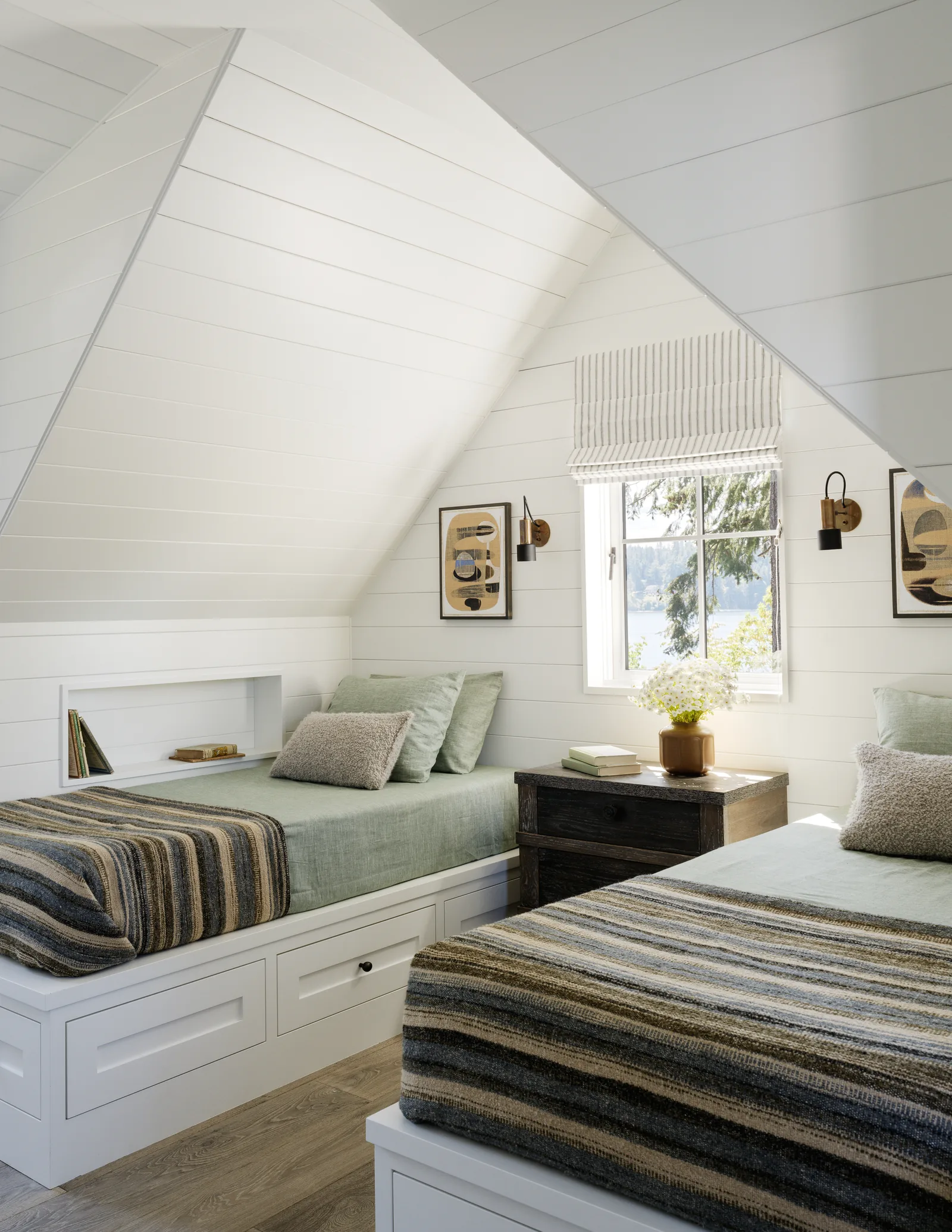 Attic bedroom showing two twin beds with built-in storage under sloped shiplap ceiling and walls, centered around a window with a striped Roman shade.