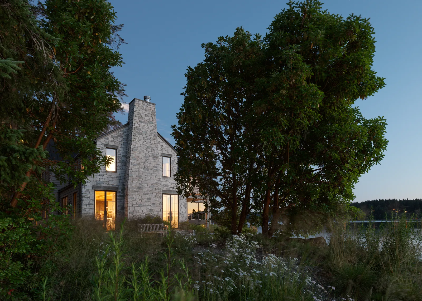 Stone house exterior with illuminated windows and visible chimney at dusk by the water.