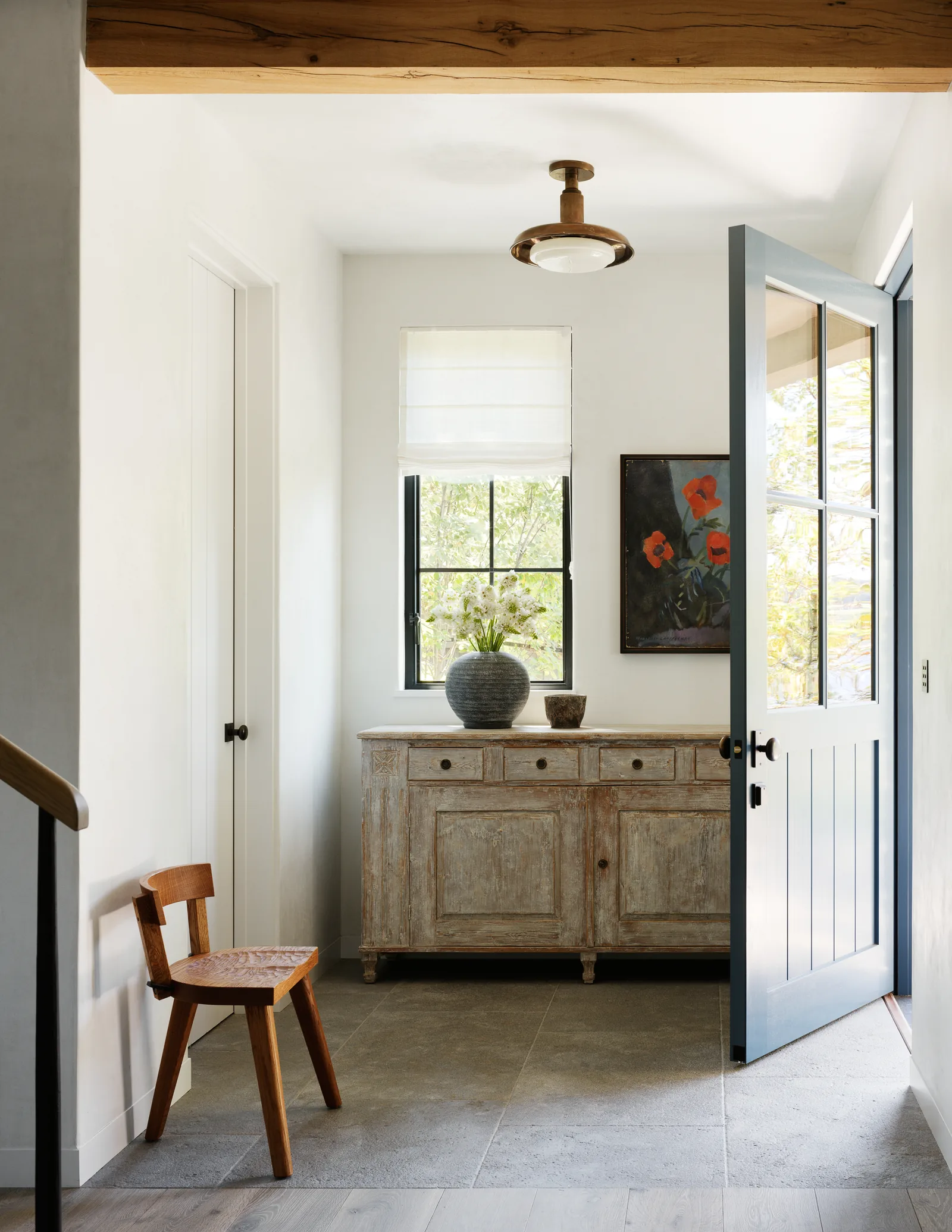 Entryway features a rustic wooden cabinet beneath a window with a roman shade and an open blue door.