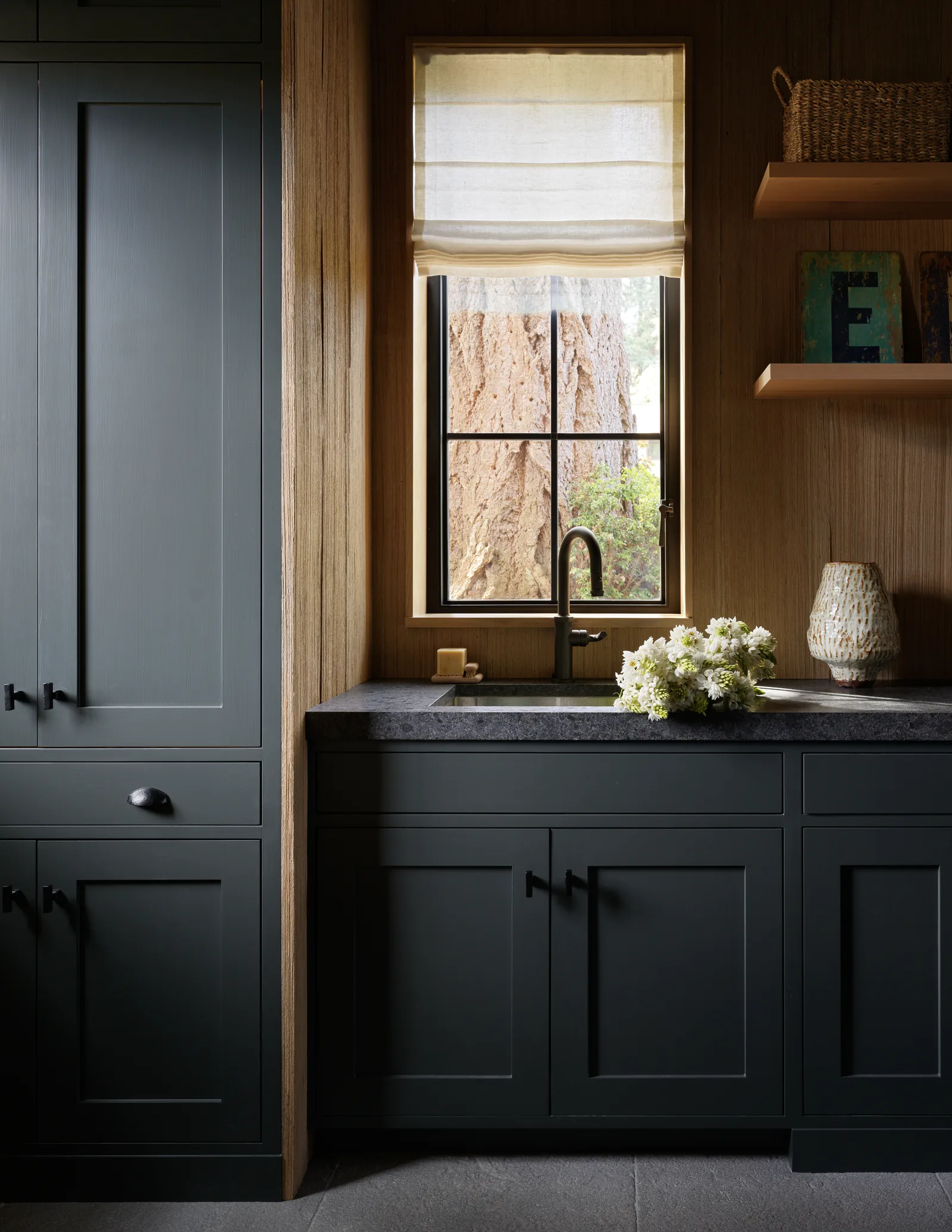 Dark cabinetry surrounds a kitchen sink beneath a window with a beige Roman shade.