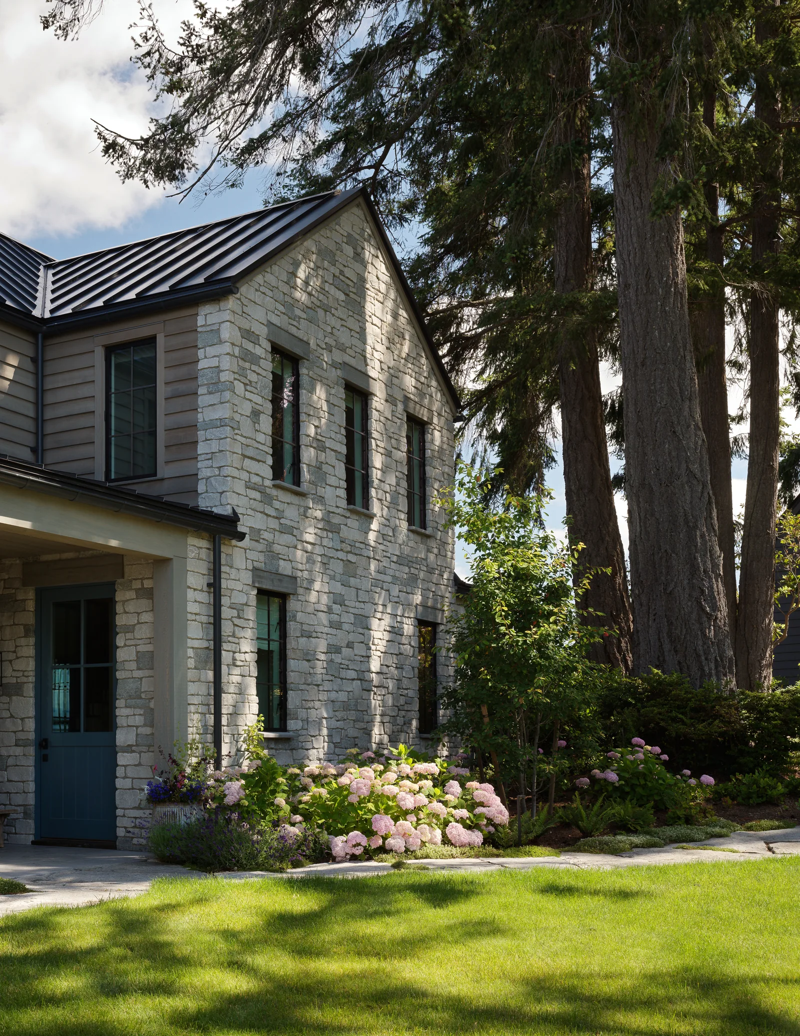Two-story stone-clad house exterior with a metal gable roof and landscaped garden bed.