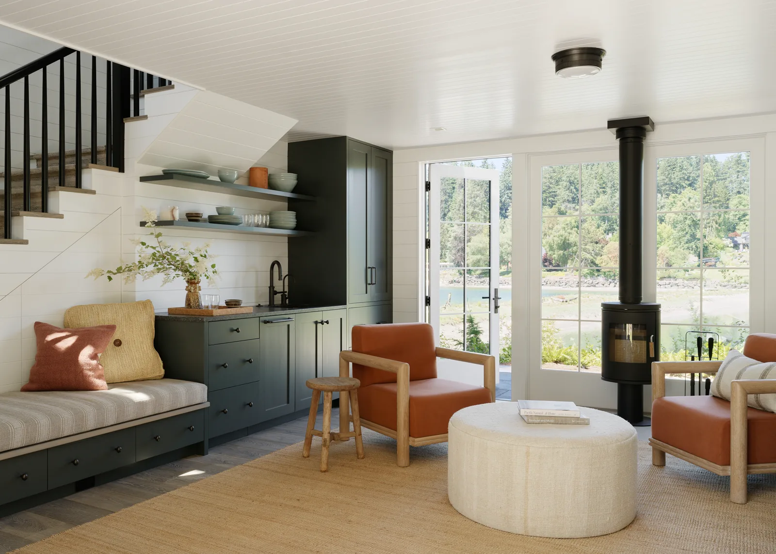 Living area features built-in bench seating under stairs, green kitchen cabinetry, freestanding wood stove, and glass French doors with water view.