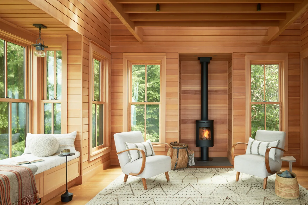 Living room of Chuckanut Studio features natural wood walls, a wood stove fireplace, and large sash windows framing forest views.