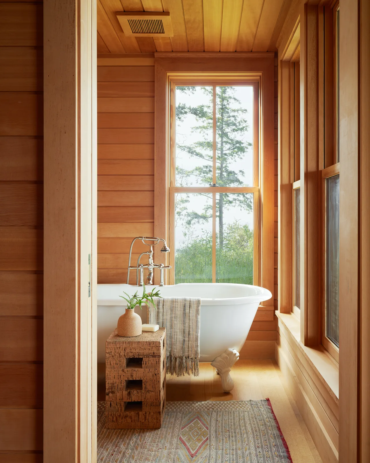 Clawfoot tub beside large window in wood-paneled bathroom with natural light and forest view.