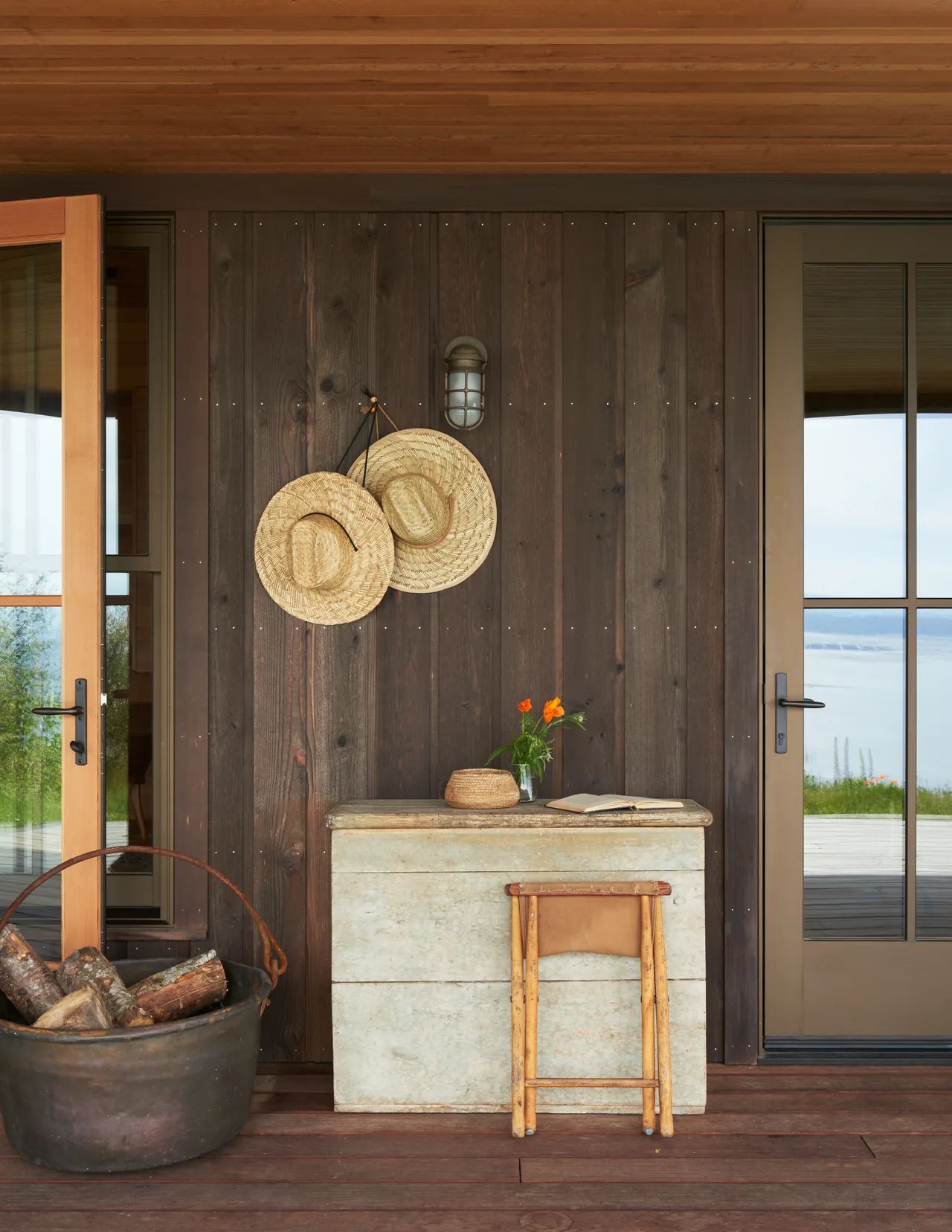 Outdoor entry with dark wood siding, decorative straw hats, weathered table and stool, and a bucket of logs beside glass doors.