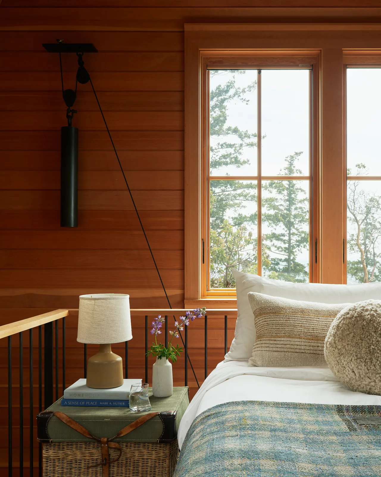 The bedroom features horizontal wood wall paneling and a window overlooking a forested area, with a bedside table holding a lamp and flowers.