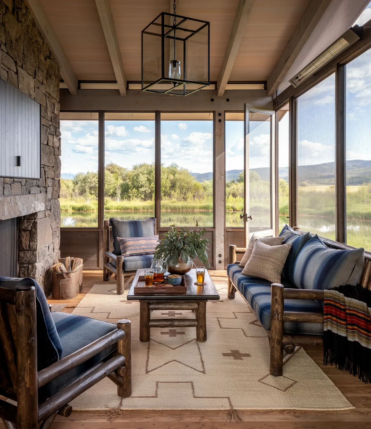 Rustic sunroom features wood-framed furniture with blue cushions, a stone fireplace, and expansive screened windows with nature views.