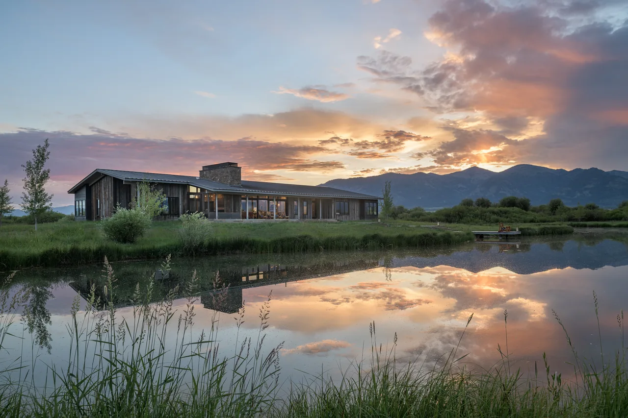 Wyoming Retreat features wide floor-to-ceiling glazing and a stone chimney with sunset reflections in a pond.