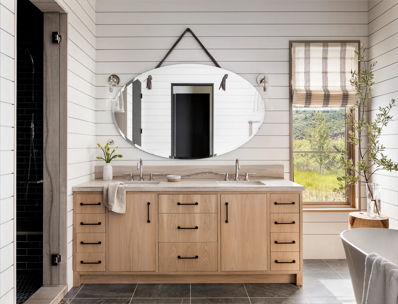 Bathroom features a double vanity with wood cabinets, an oval mirror hung by leather straps, and a window with a striped Roman shade overlooking landscape.