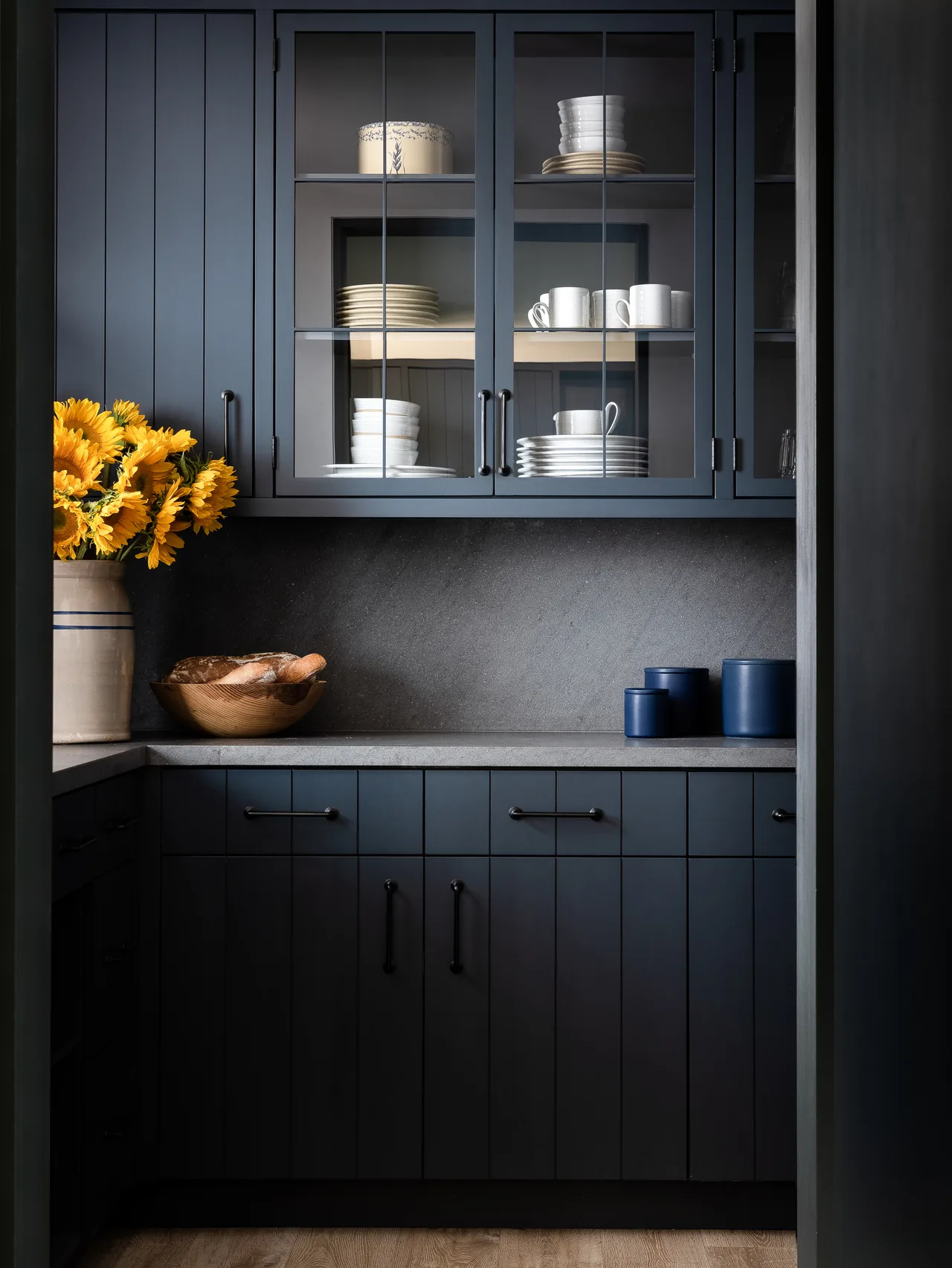 Dark blue kitchen cabinets with glass doors show neatly stacked dishes above a counter with sunflowers and a bread bowl.