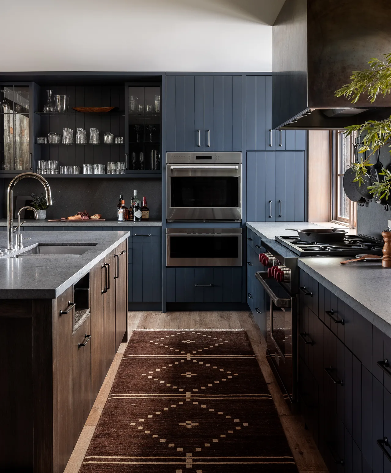 The Wyoming Retreat kitchen features blue cabinetry, a wooden island, stainless steel double ovens, and a patterned brown rug on wood flooring.