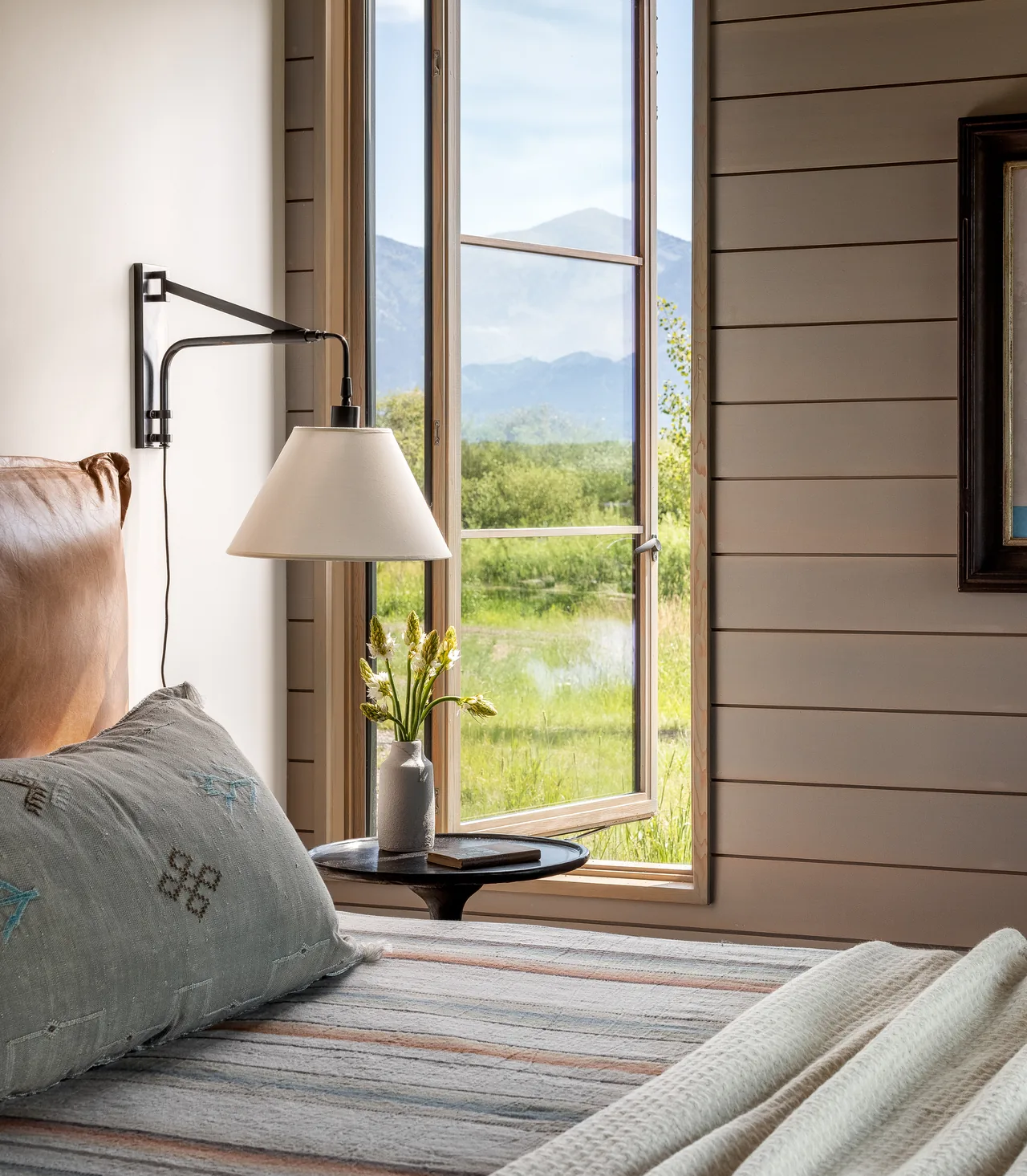 Bedroom interior with a leather headboard, wall-mounted lamp, and window revealing a mountain view.