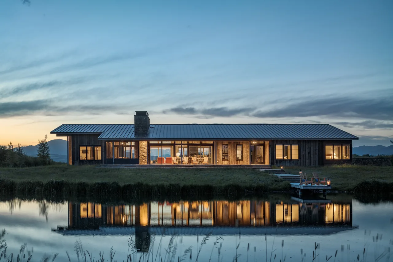 Wyoming Retreat features a gable roof, stone chimney, and extensive floor-to-ceiling glazing with black frames, seen at evening reflected in water.