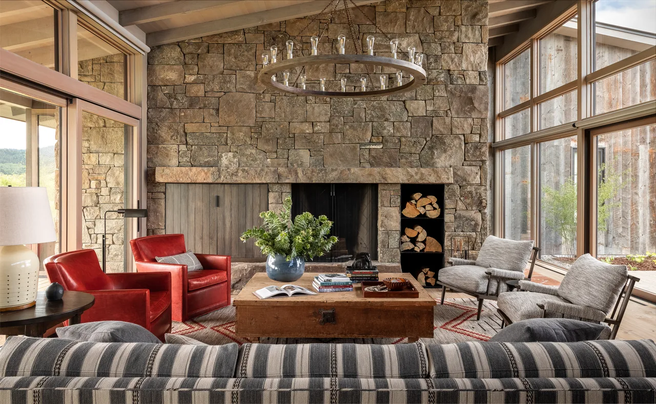 Wyoming Retreat living room features a large stone fireplace with firewood storage and floor-to-ceiling windows overlooking the outdoor landscape.